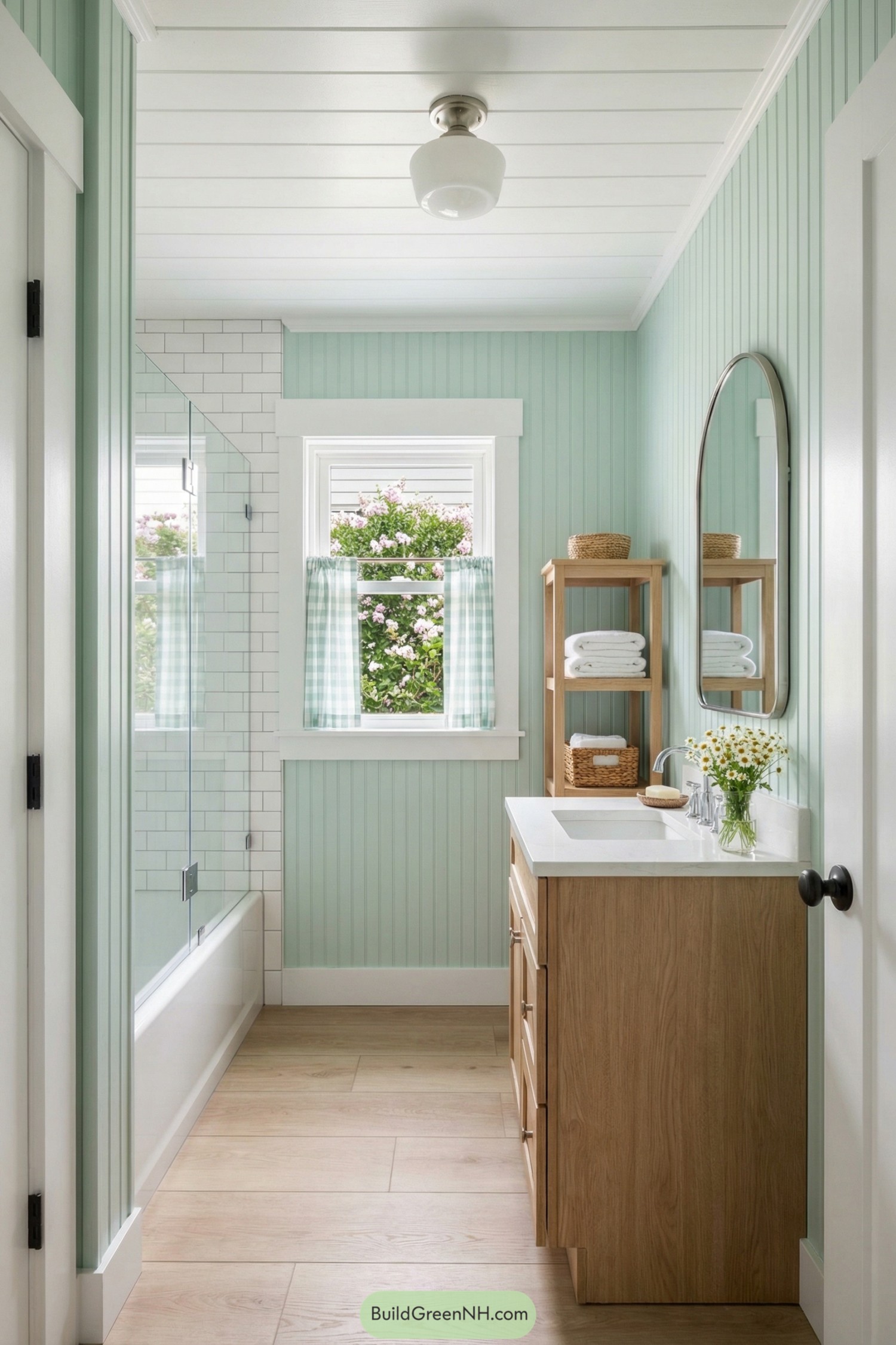 Small mint green beadboard bathroom with wood vanity, glass tub shower, and window overlooking blooming garden