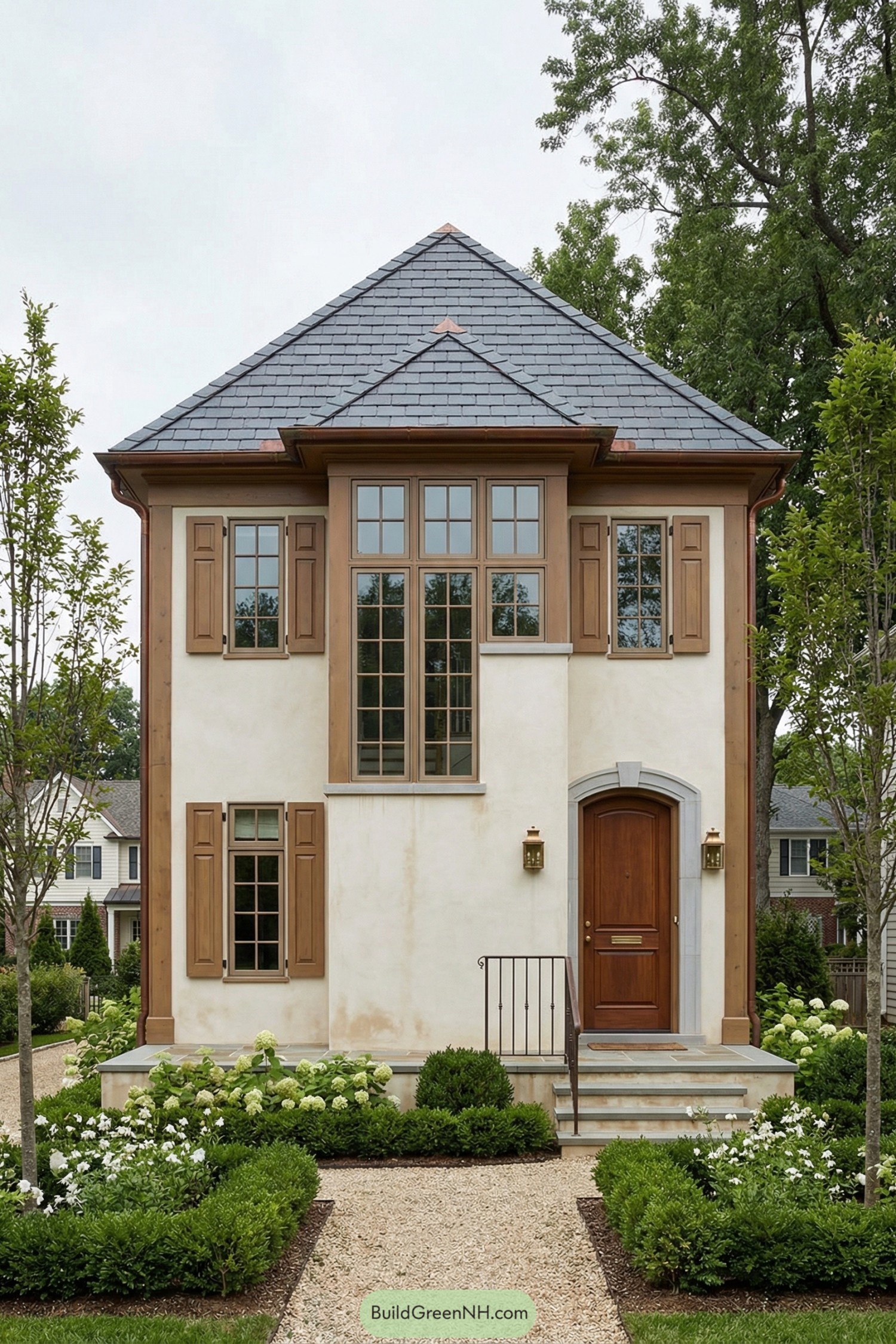 Compact cream stucco cottage with tall slate roof, wood shutters, and manicured front garden