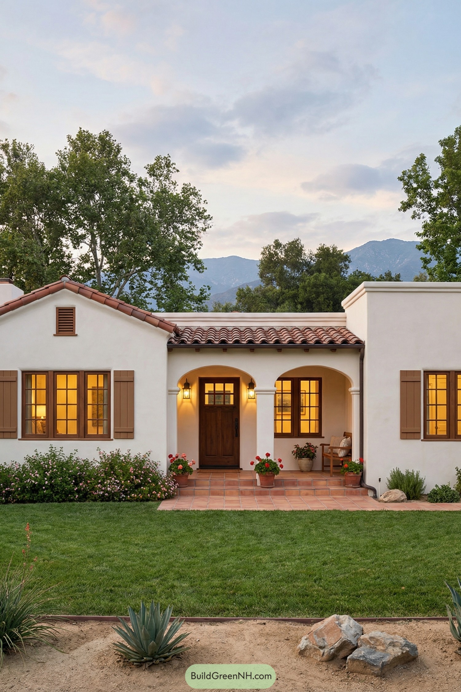 Single story white stucco Spanish style house with red tile roof arches and wooden shutters, set in a green yard with potted flowers
