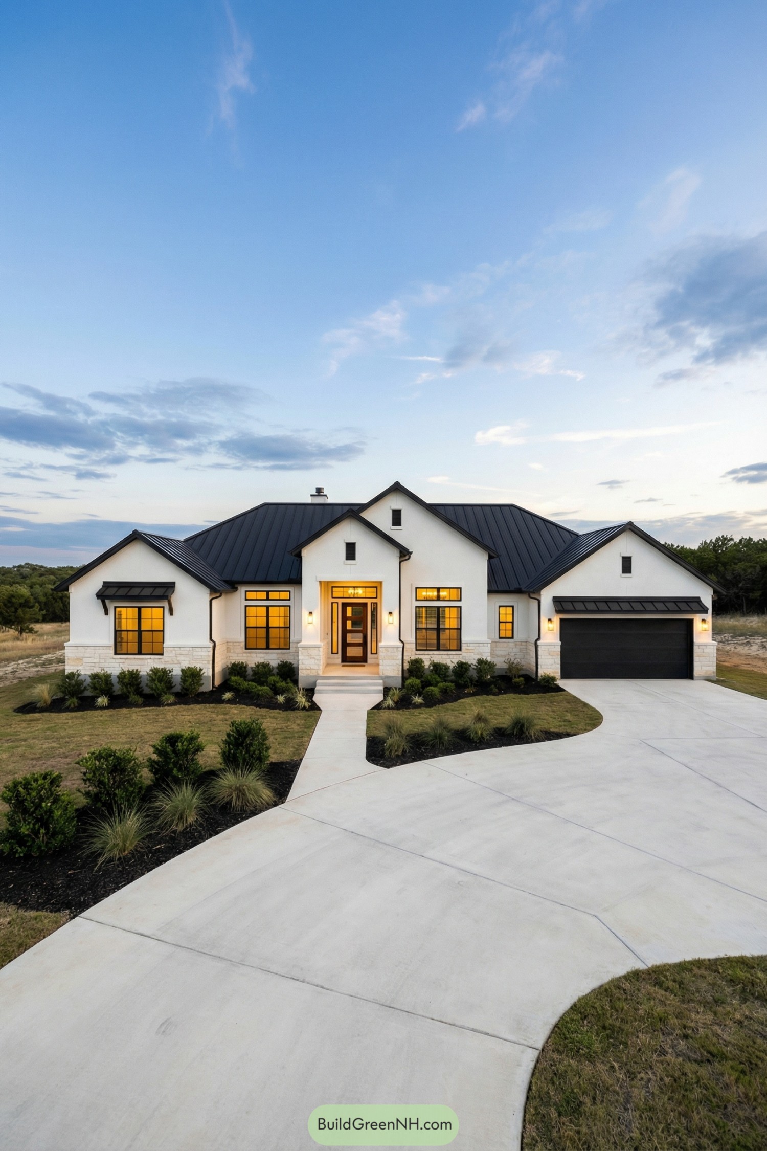 Single story white house with black metal roof, warm lit windows, and curved driveway framed by simple landscaping