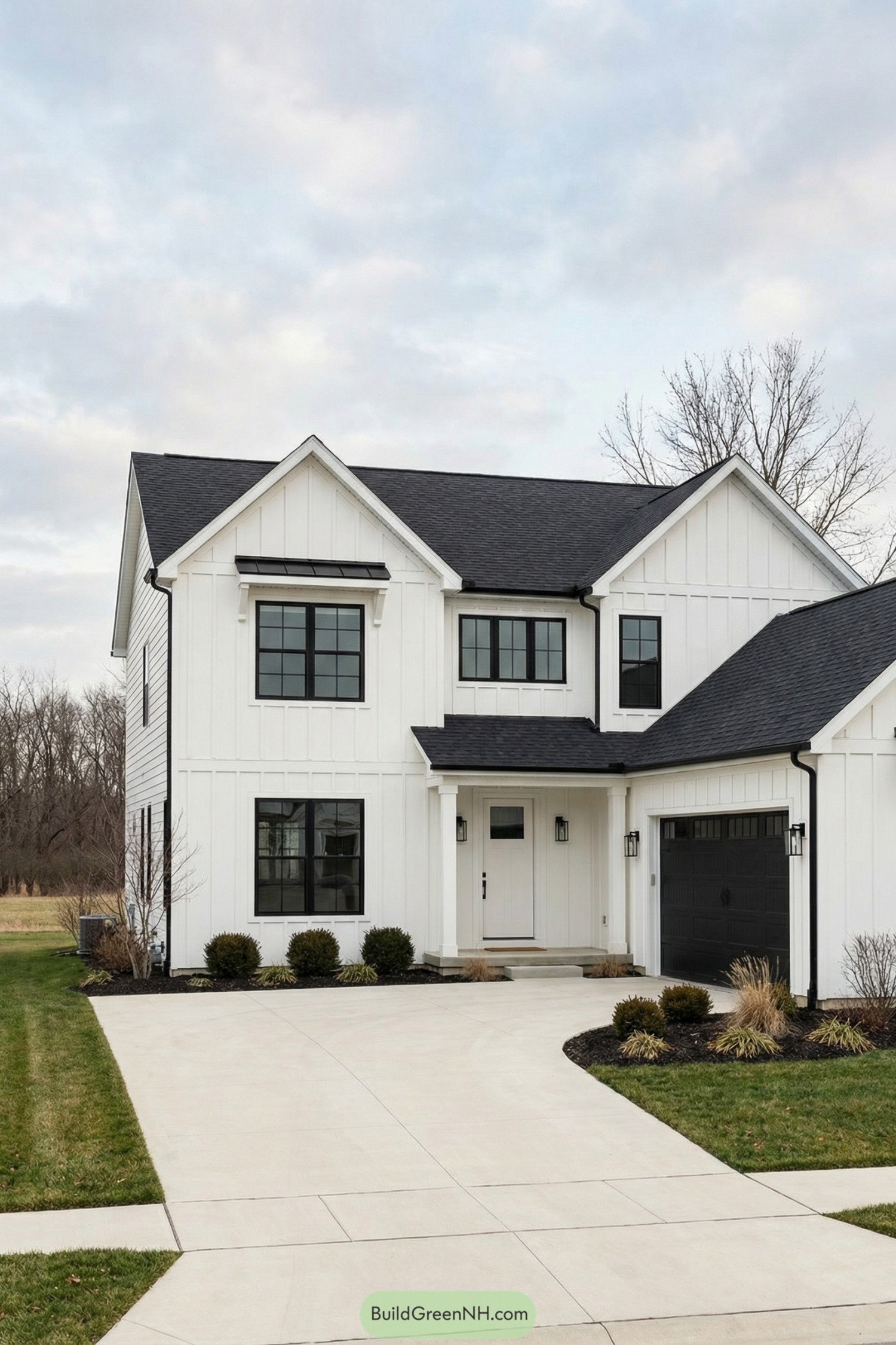 high-res photo of white house black windows, modern farmhouse facade with clean lines and cross-gabled front, crisp white vertical board-and-batten siding with minimal black trim, asymmetrical two-story massing with attached single-story garage wing, black asphalt shingle roof with moderate pitch and simple eaves, black framed grid-style windows evenly spaced on both levels, black double garage door with horizontal panels, simple white front door recessed under small covered porch with square white columns and black wall sconces, smooth light concrete driveway leading to garage and entry, low dark mulch beds with compact shrubs and ornamental grasses along foundation, neatly edged green lawn bordering driveway and walkway, sparse leafless trees and shrubs around property, soft cloudy sky and bare tree line in the distant background creating a calm suburban setting, single real-life photo, high-resolution, architectural photography, soft lighting, cinematic composition, strictly no collages.