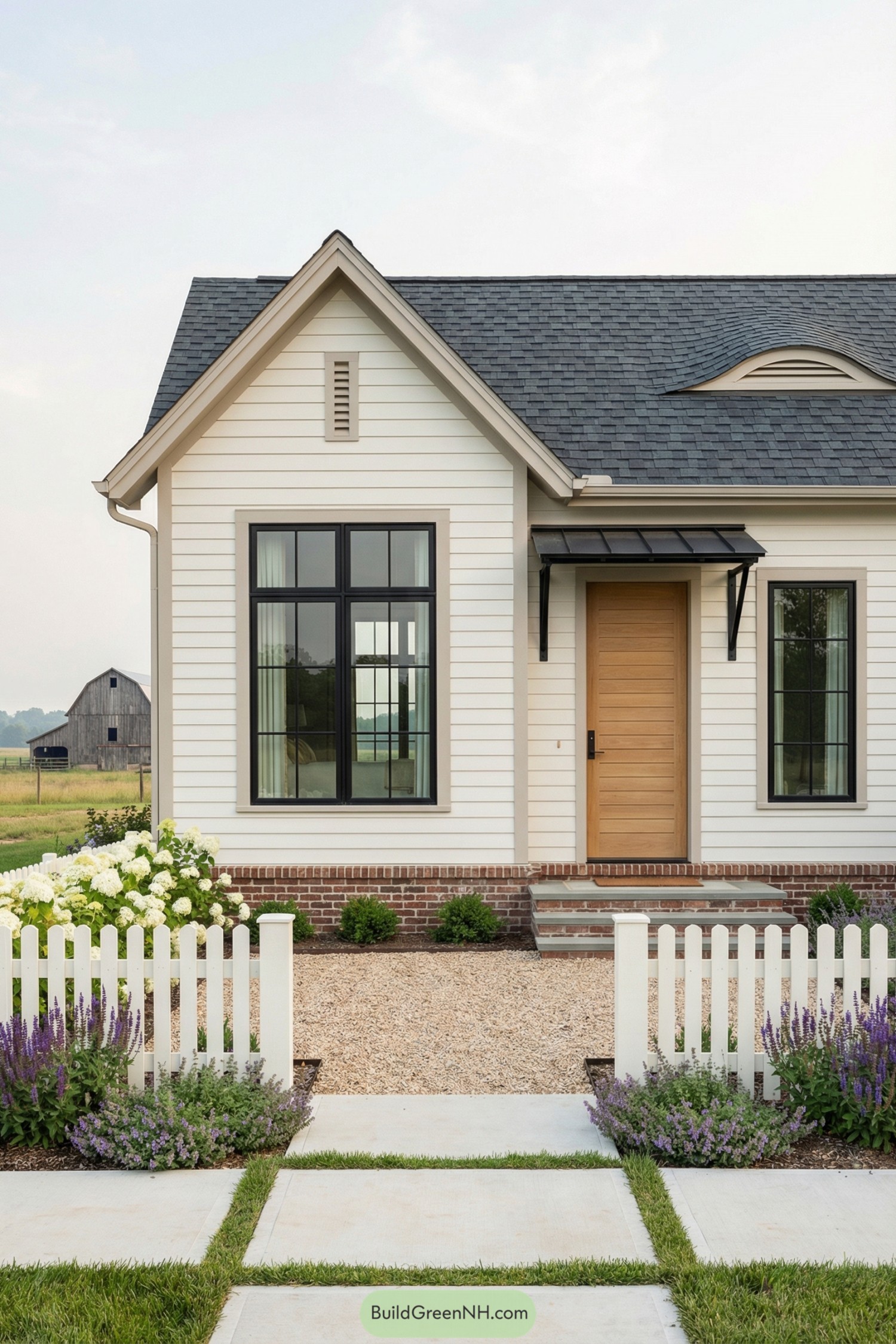 White cottage with black windows and a wood front door behind a picket fence and flower beds
