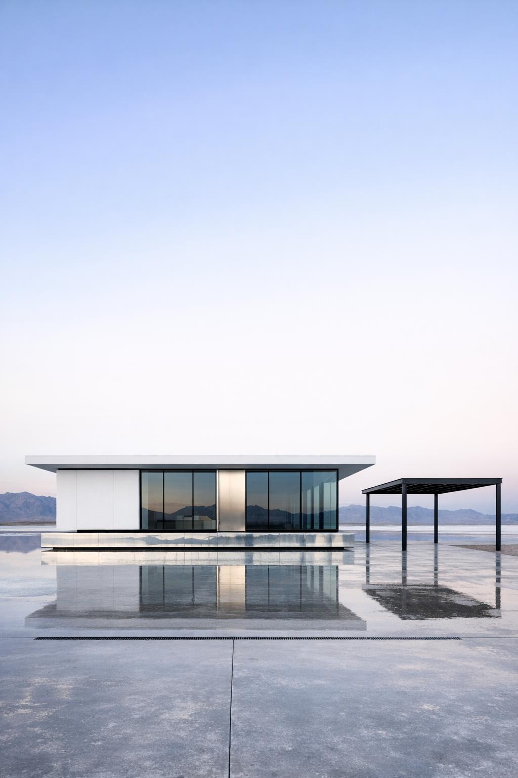 Minimalist white house with glass walls reflected on a smooth concrete surface in an open landscape