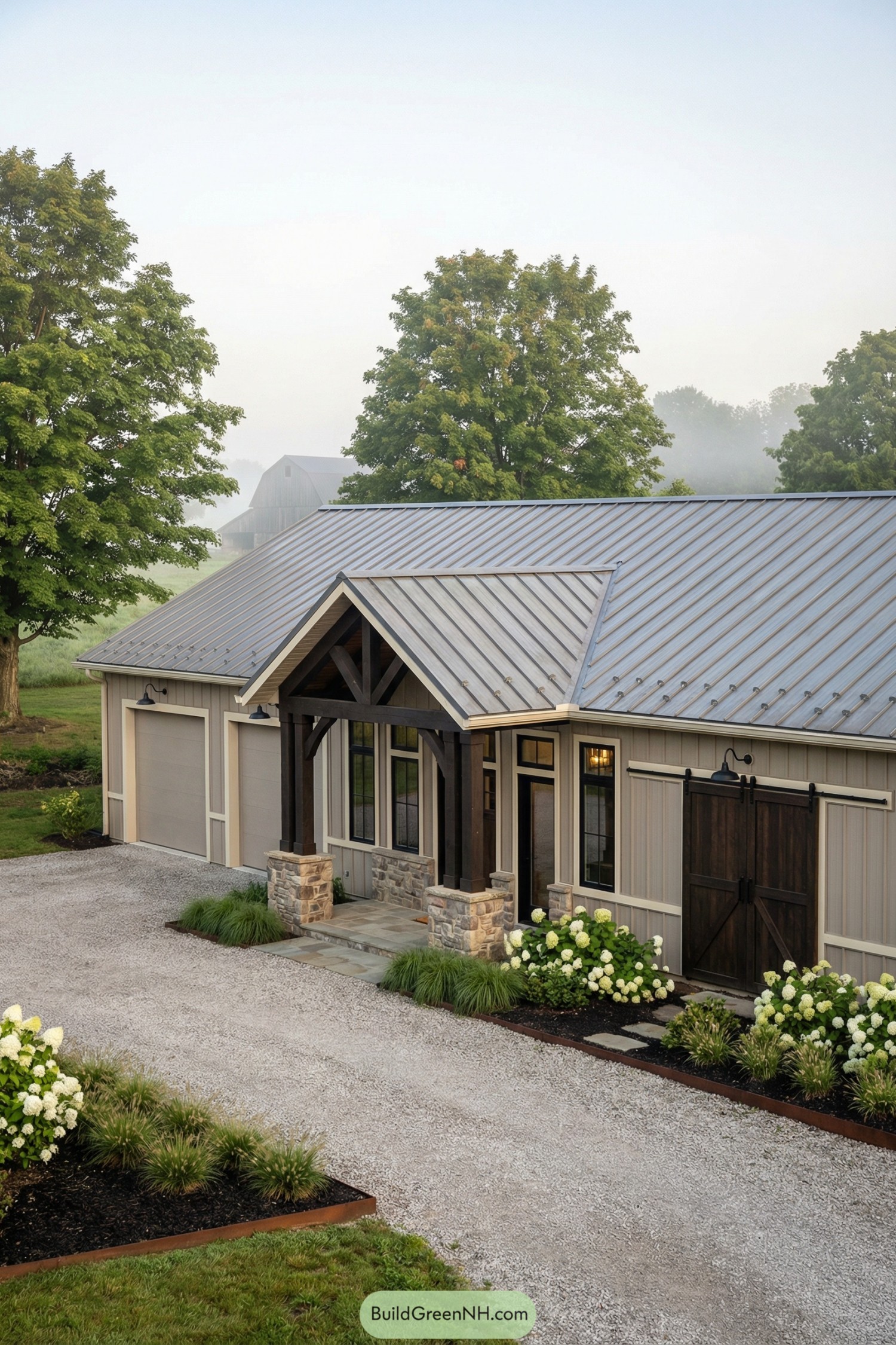 A ranch-style barndominium with a refined rustic facade, defined by a central gable porch with exposed trusses and two flanking garage-like barn bays expressed as simple volumes; colors of light taupe metal siding, creamy trim, and dark espresso wood; a wide single-story composition with a stepped facade depth; materials include ribbed steel panels, natural fieldstone piers, and heavy timber brackets; a medium-pitch gable roof in aged zinc standing seam with snow guards; tall rectangular windows with divided-light upper sections in black frames; a pair of sliding barn-style doors in stained wood on black tracks and a separate glazed entry door to the side; outdoor structures include a crushed-limestone drive, black gooseneck sconces, and a covered front stoop with stone paving; landscaping of hydrangea masses, low ornamental grasses, and mulched beds edged in steel; environment of a lush rural setting with mature maples and a distant barn silhouette; soft morning mist and diffuse light for a calm, cinematic mood, single real-life photo, high-resolution, architectural photography, soft lighting, cinematic composition, strictly no collages.
