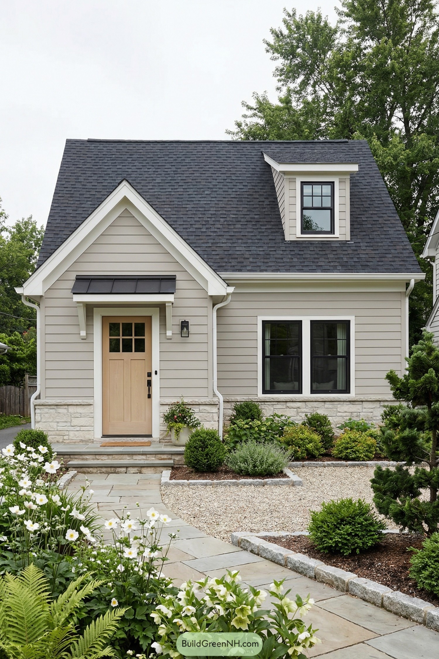 Compact gray cottage with steep roof, light wood front door, dormer window, and tidy flower lined walkway