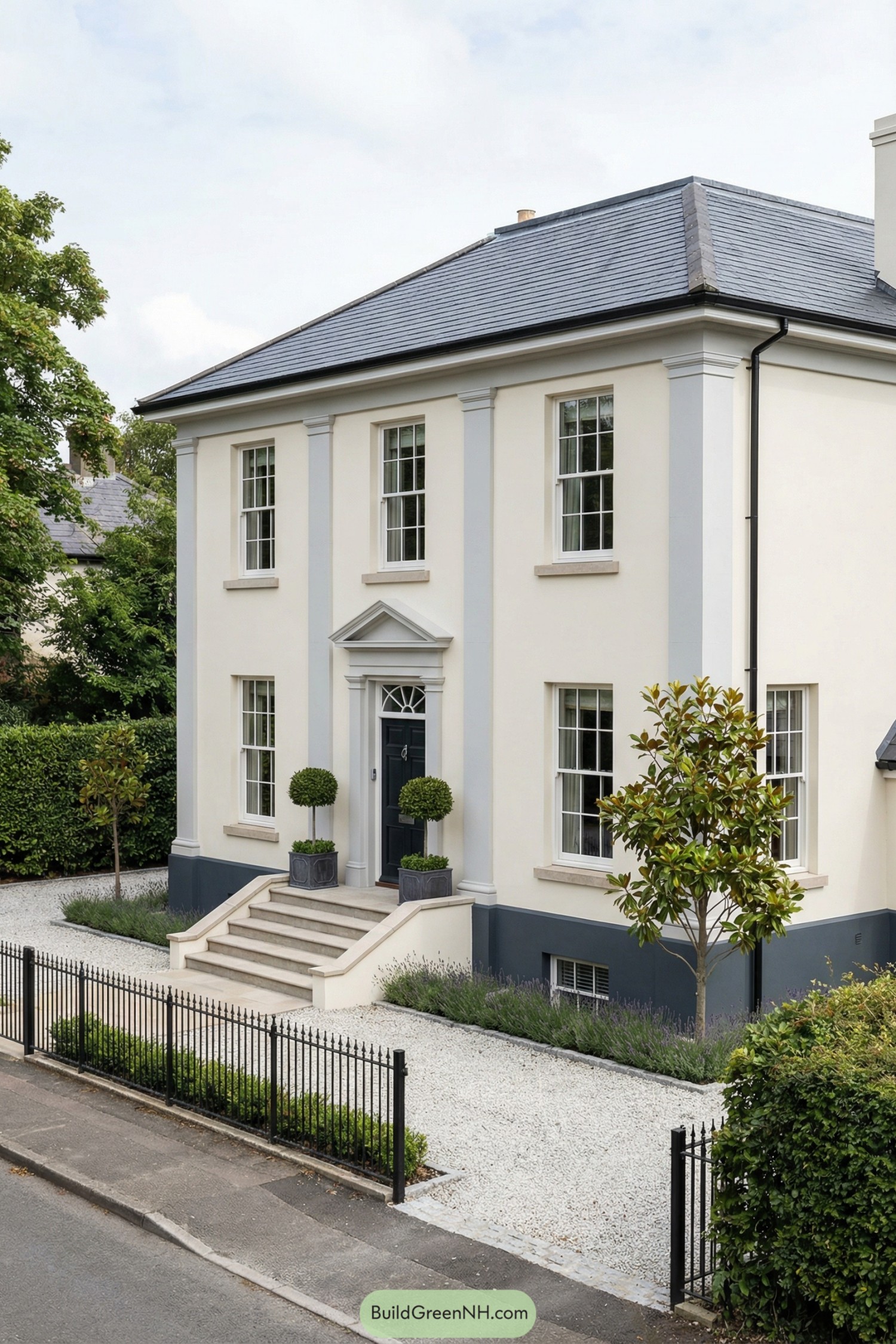 Cream colored two story Georgian style house with dark roof, front steps, and neat gravel garden with low plants and small trees