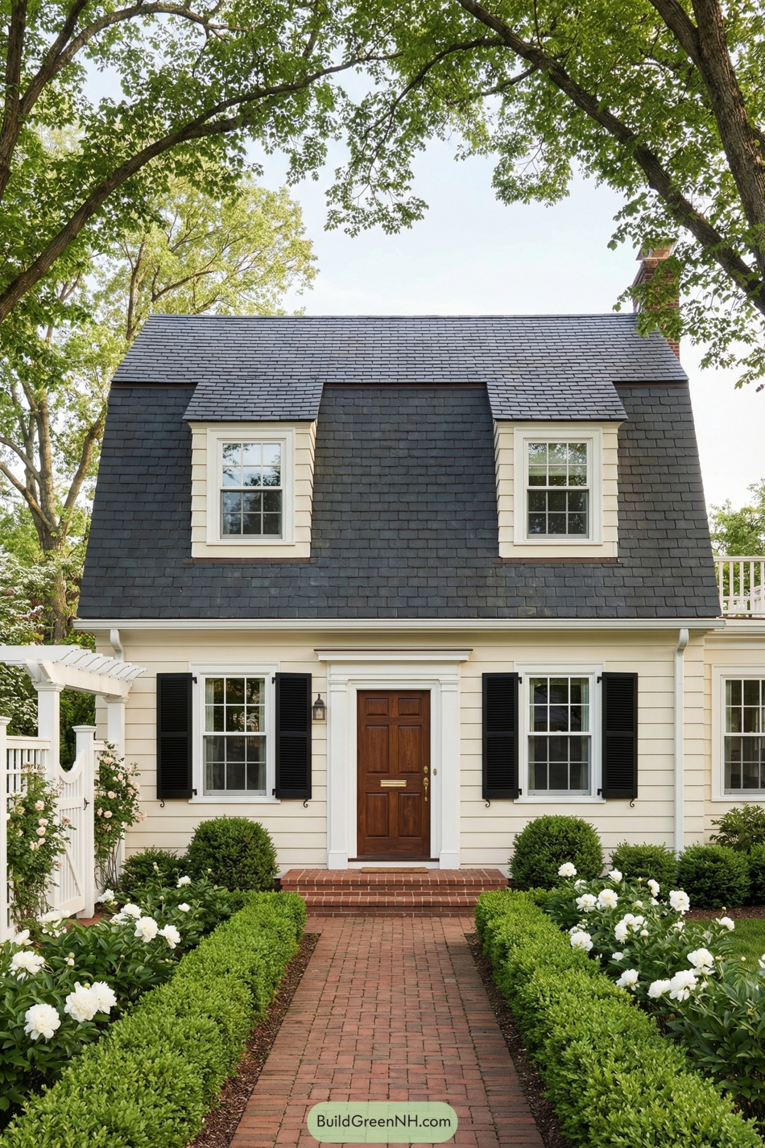 Small cream Dutch Colonial style house with gambrel roof, brick path, and lush front garden