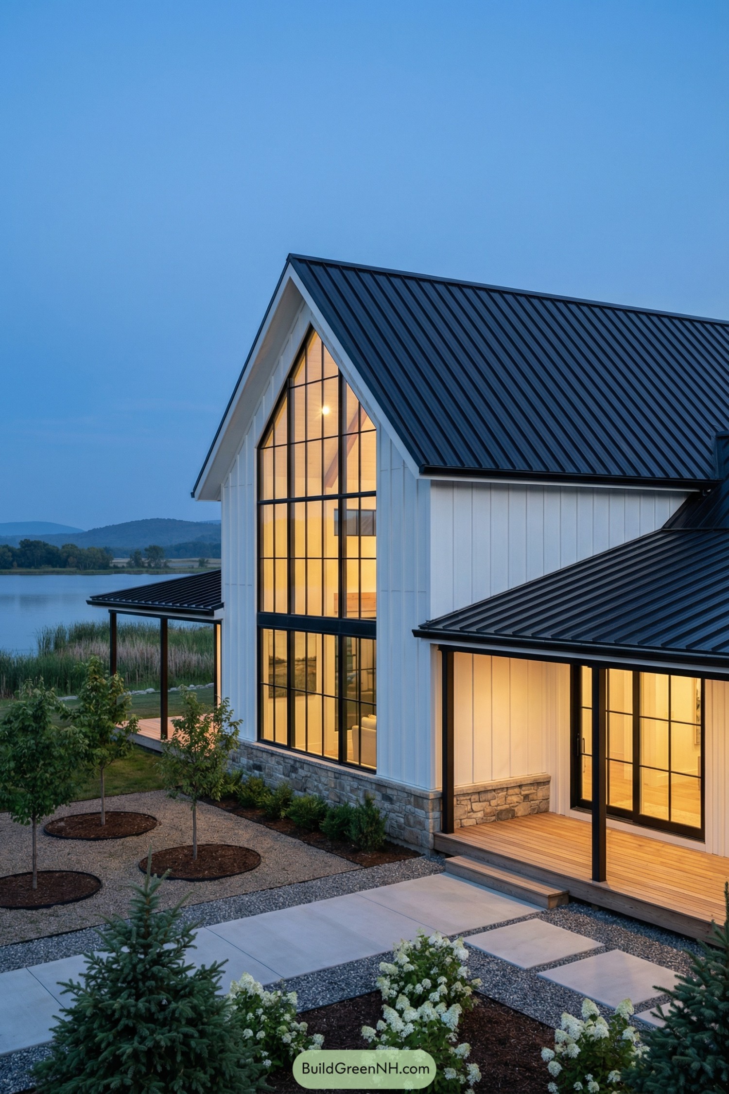 Modern white barndominium with tall black windows by a lake at dusk