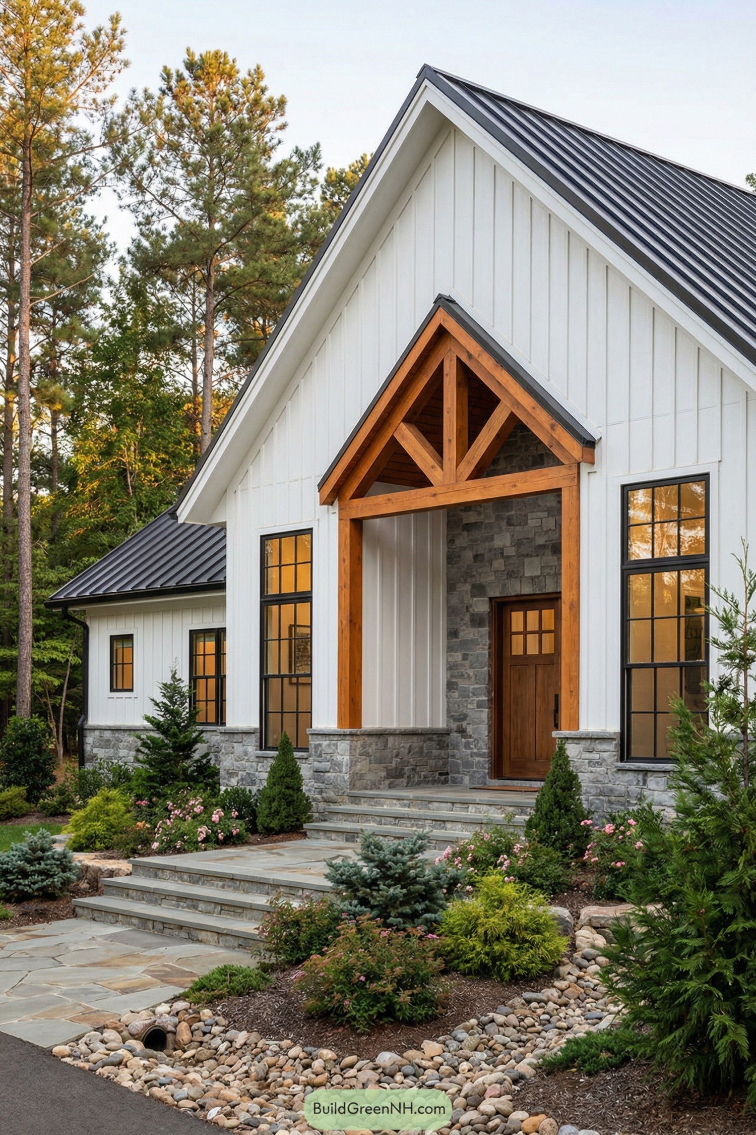 Modern farmhouse with white board and batten siding, stone base, black metal roof, and a warm timber framed entry surrounded by lush landscaping