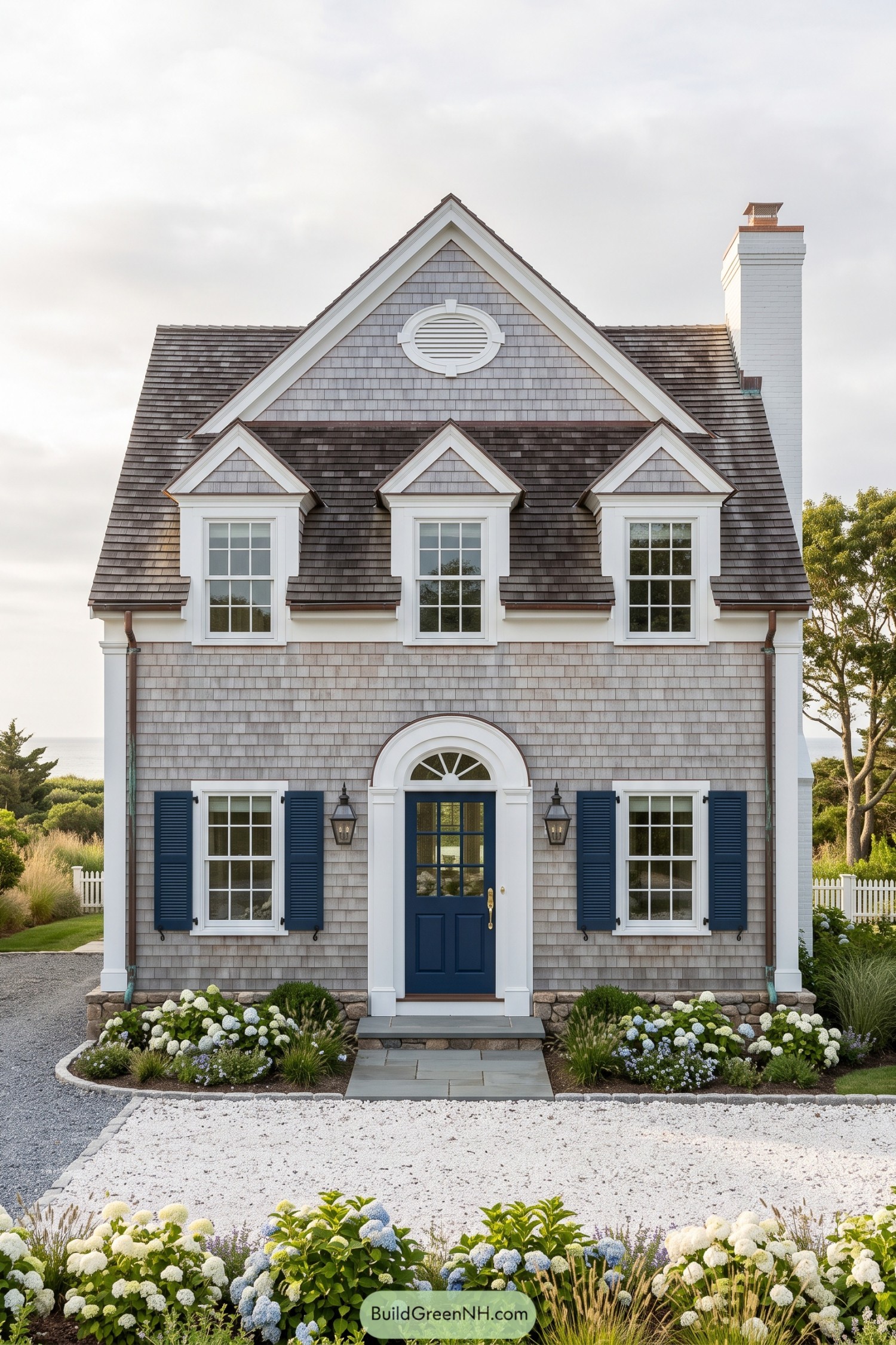 Gray shingled Cape Cod house with blue shutters three dormers and an arched entry framed by hydrangeas