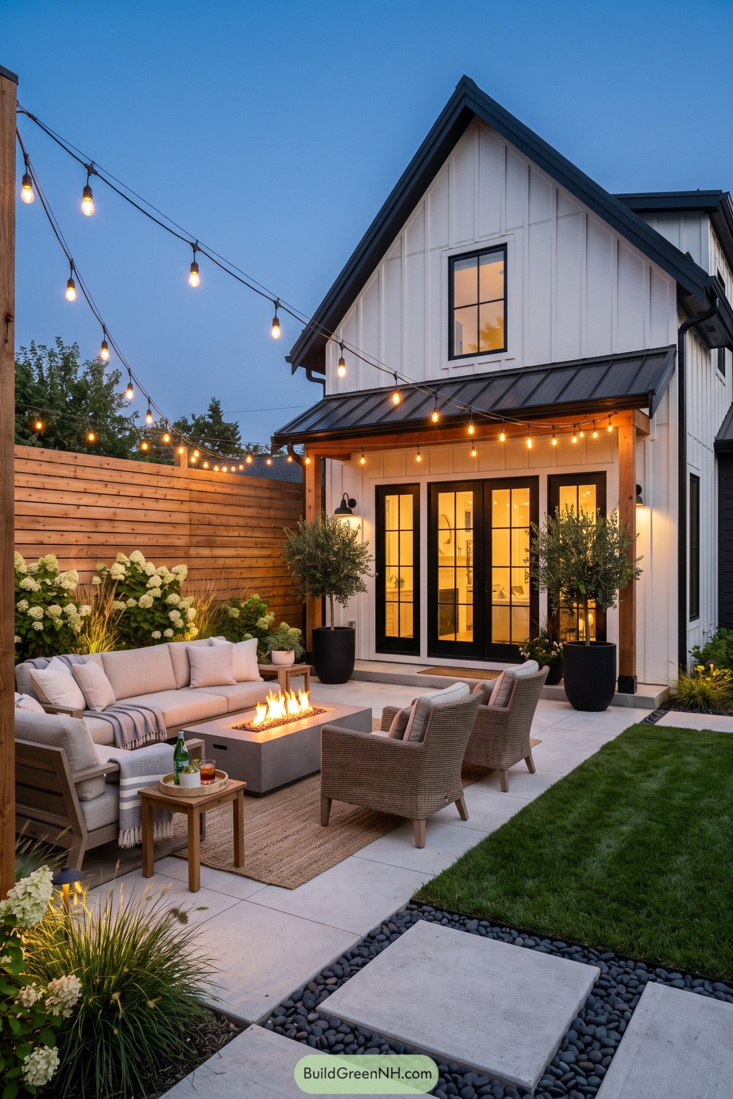 Cushioned seating surrounds a rectangular fire table on a backyard patio with cedar fencing and overhead string bulbs